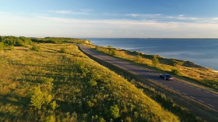 Aerial view of driving black car on the road along the cliff near the sea in summer at sunset