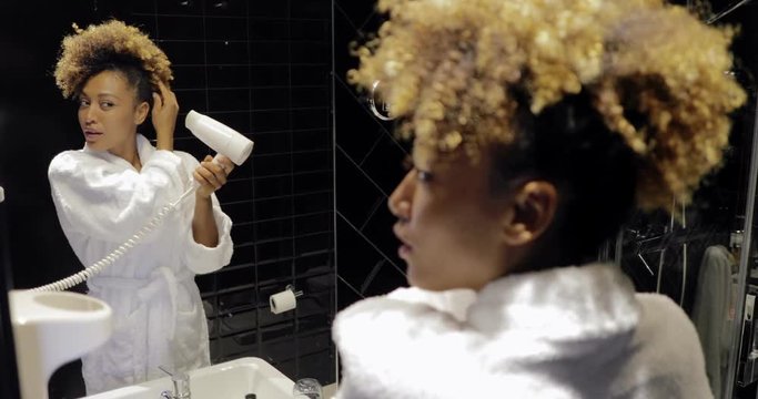 Confident young woman in soft bathrobe styling curly hair with hairdryer while posing in front of mirror in hotel bathroom.