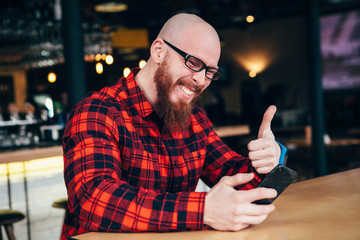 Handsome bearded man in casual clothes sitting at coffee shot with phone making video call to friend. Caucasian male having video chat on smart phone at cafe.