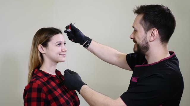 Portrait of happy female client and male beautician in healthcare clinic cabinet of beauty salon. Male cosmetologist in black gloves making permanent makeup procedure on female eyebrows. Thums up