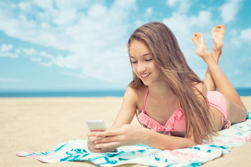 Pretty teenager girl using a smart phone lying on the beach with the sea and horizon in the background