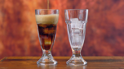 Pouring cola soft drink on ice in tall cafe glasses against a rustic wood background.