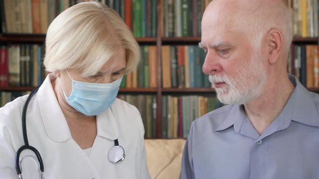 Female professional doctor in white coat and medical mask at work. Senior woman physician talking to sick senior male patient at home consulting about new drug. Showing bottle of pills capsules