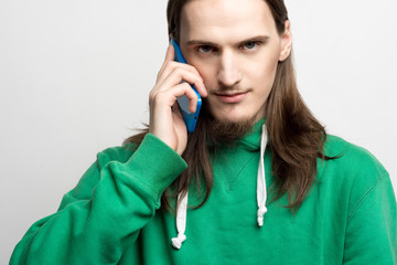 Portrait of young handsome man in green hoodie looking at the camera, talking on cell phone agains...
