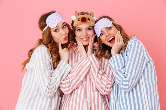 Three beautiful young girls 20s wearing colorful striped pyjamas and sleeping masks having fun during girlish sleepover, isolated over pink background