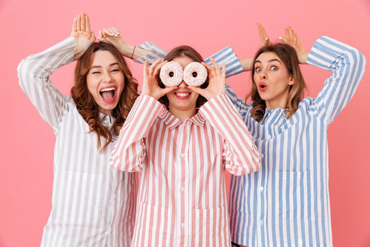 Image of happy and young women 20s with brown hair wearing leisure clothings having fun with sweet desserts donuts at slumber party, isolated over pink background