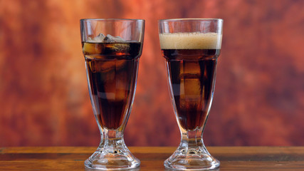 Pouring cola soft drink on ice in tall cafe glasses against a rustic wood background.