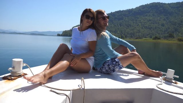 Young pair sitting back to back and posing on bow of boat at sunny day. Happy couple in love spending time together on deck of ship and enjoying summer travel. Concept of vacation or holiday. Close up