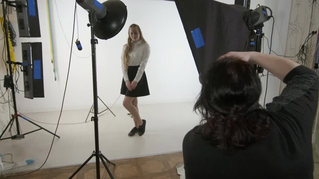 Studio photography. Teen girl poses for photographer in photo studio. Woman photographing young blonde model on white background. Backstage shot during indoor photoshoot.