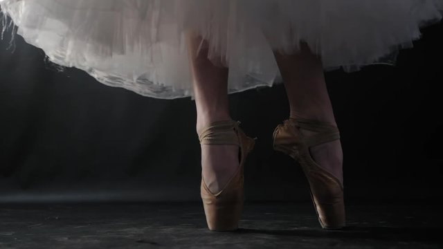 Close up of ballet dancer as she practices exercises on dark stage or studio. Woman's feet in pointe shoes. Ballerina shows classic ballet pas. Slow motion. Flare, gimbal shot.