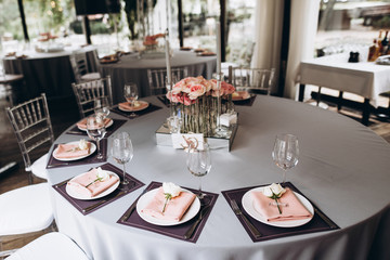 Wedding banquet table which is beautifully decorated in pastel colors, with white roses on plates, and a beautiful composition of roses and candles in the center of the table