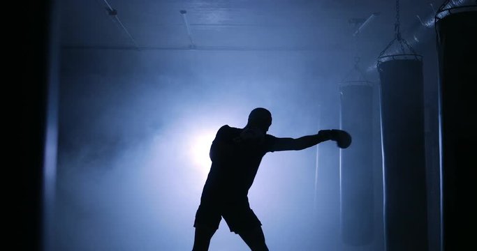 Strong male boxer training in a dark smoky room. Silhouette on a dark background