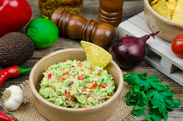 A bowl of delicious homemade guacamole next to fresh ingredients on a table served with tortilla chips