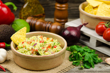 A bowl of delicious homemade guacamole next to fresh ingredients on a table served with tortilla chips