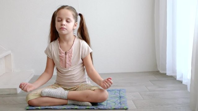 little cute girl sitting in yoga lotus pose and meditates near the window