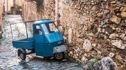 Fototapeta na wymiar Castelmola, Taormina, Messina. March 2017: traditional vintage vehicle with three weels and blue metal paint, called Ape car, parked on the street, next to a rural house with big stone wall.