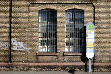 Bushaltestelle an einer stillgelegten Fabrik / Ein Bushaltestellezeichen steht vor der Backsteinwand eines stillgelegten Industriegeländes einer alten Fabrik mit einem vergitterten Fenster.