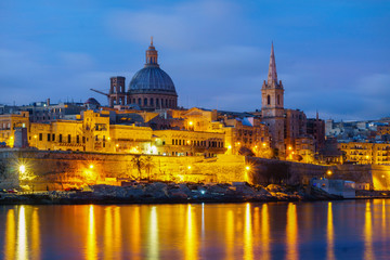 Obraz na płótnie Canvas Night view of Valletta, capital of Malta. Beautiful spires and St. Pauls Cathedral and Charmelite Church skyline under blue night sky, lights reflected in the sea bay.