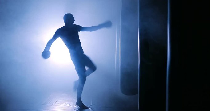 Agressive kickboxing man punching a boxing bag with boxing gloves in a smoky gym. Silhouette on a dark background