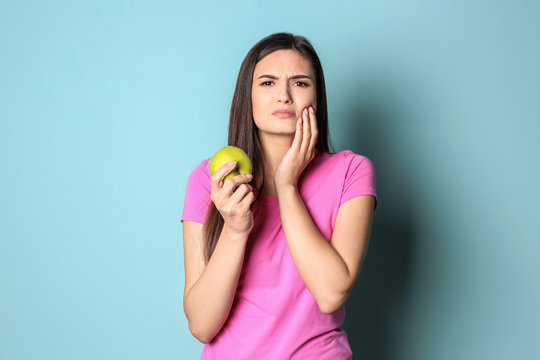 Young woman with sensitive teeth and apple on color background