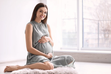 Young pregnant woman sitting on floor near window at home