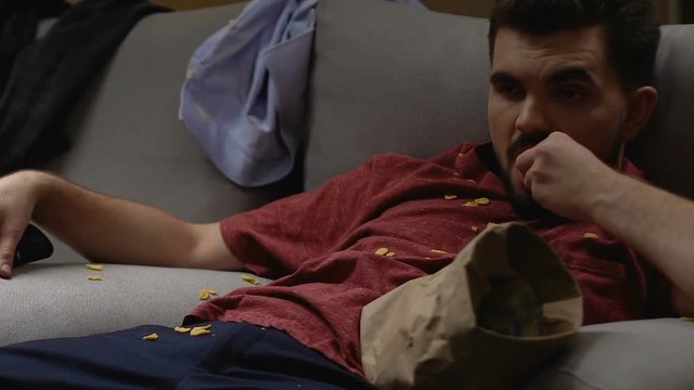 Unemployed bachelor watching TV lying on sofa, burning his life in garbage