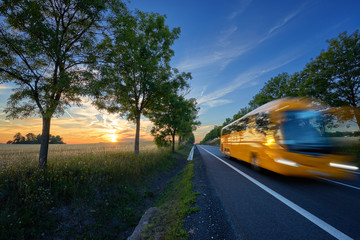 Motion blurred yellow bus traveling on the road in the avenue of trees in a rural landscape at sunset © am