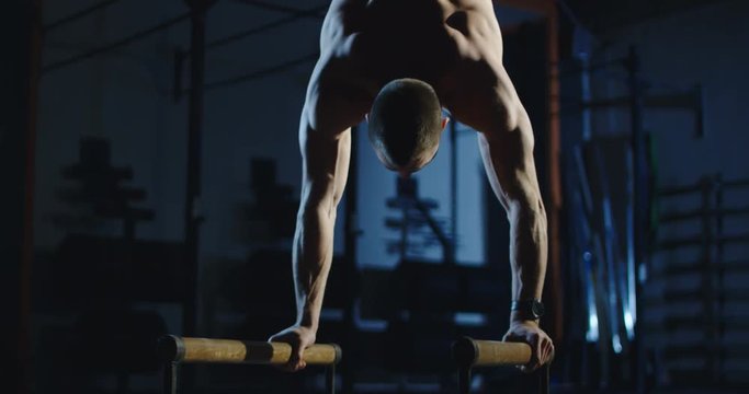 Muscular shirtless man working out on bars training gymnastics in dark gym.