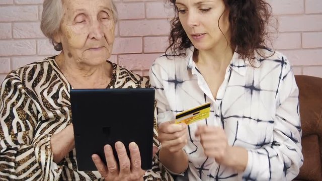 An elderly with a credit card. Elderly pays. A young woman is teaching an old woman to use a credit card and tablet.