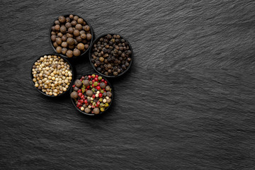 Fototapeta na wymiar Black, Green, Pink, White and Fragrant Pepper Grains in Round Jars Placed on Black Stone Surface Background