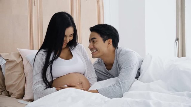 Young husband touching belly of smiling pregnant wife on bed