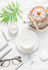 Flat lay cozy morning inspiration planning female table - teapot, tea cup, notepad, glasses, lipstick, face cream, green leaf flower on light background, top view