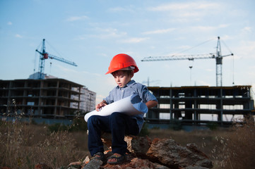 handsome little boy wearing orange helmet sitting on construction site with paper plan in hands on crane and building background
