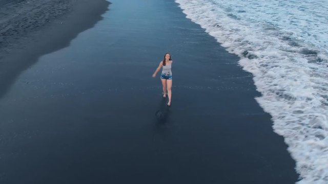 Aerial footage of the ocean beach with black sand and waves at sunset on Bali with beautiful happy young walking female model in jeans shorts and a gray T-shirt, video 4K