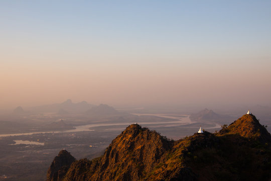 Scenic sunset view of white ancient buddhist stupas on peaks of textured mountains in yellow sun light. Misty valley with winding river and shadowy mountains background. Kayin State, Myanmar.