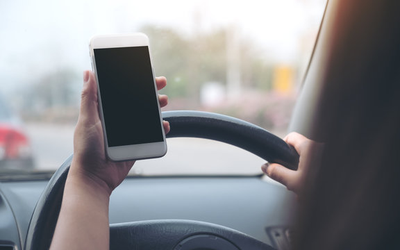 Mockup image of a woman's hand holding and using white mobile phone with blank black desktop screen while driving car on the road
