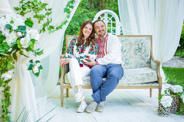 Obraz premium Beautiful young couple in Ukrainian national shirts embroidered shirts sitting on the couch in the background white gazebo in the park