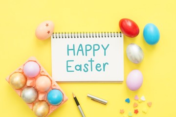 Flat lay top view colorful easter egg painted in pastel colors composition and text Happy Easter Day on notebook with paint brush on yellow pastel color background. Top view Easter day background.