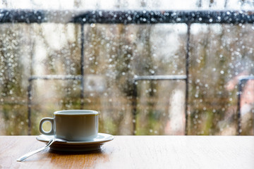 A white espresso cup with a spoon on a wooden table in front of a glass window covered with rain drops.