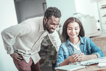 Young people. Positive nice joyful man standing behind his friend and looking at her smartphone while being in a positive mood