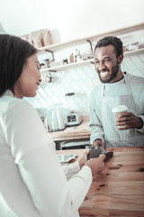 Obraz premium Friendly barista. Cheerful positive delighted man smiling and talking to the customer while giving her coffee