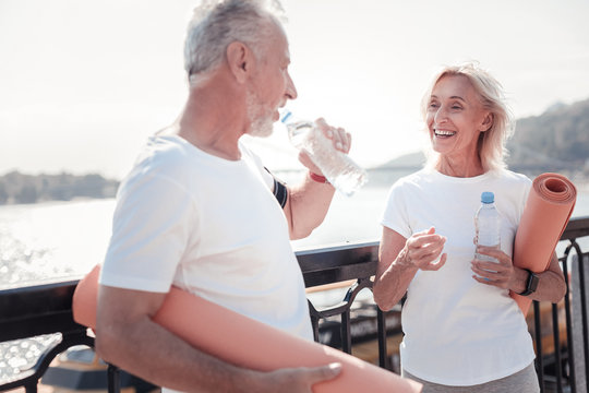 Good tome together. Happy senior sporting woman standing opposite man preparing for the training and drinking water.