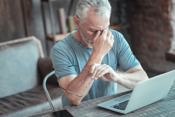 Need to relax. Exhausted senior occupied man sitting by the table opposite the laptop holding the glasses and having head ache.