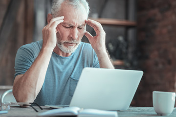 Its so difficult. Serious sick unshaken man sitting in the room by the table closing eyes and having head ache.