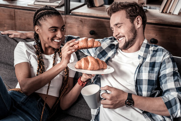 Tasty croissant. Cheerful positive young woman holding a croissant and feeding her boyfriend while having breakfast with him
