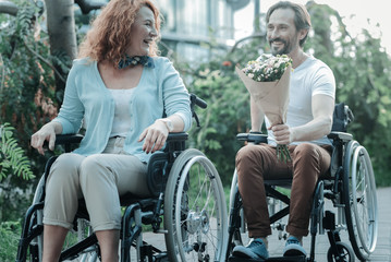 Wait me please. Handsome man keeping smile on his face and holding bunch of flowers while being on wheelchair