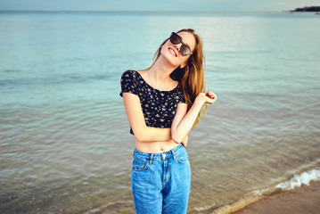 Stylish woman with long hair standing in blouse and jeans shorts. Beautiful girl walking at the beach at sunset