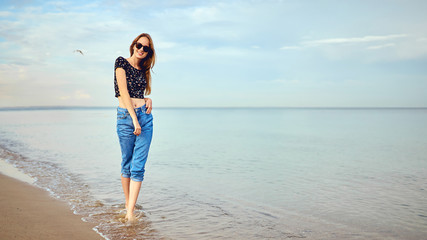 Stylish woman with long hair standing in blouse and jeans shorts. Beautiful girl walking at the beach at sunset