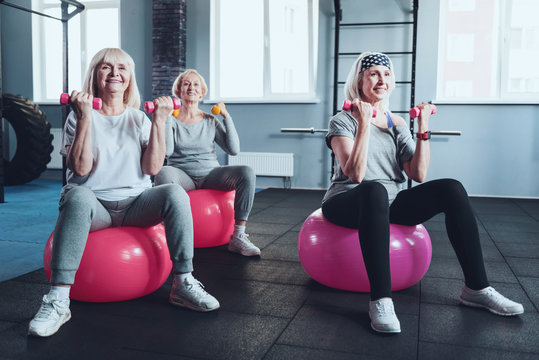 Full Of Energy And Joy. Focused Senior Female Friends Smiling While Taking An Exercise Class And Training With Dumbbells While Sitting On Fitness Ball In A Gym.