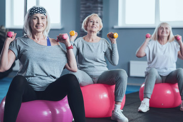 Stronger body and mental health. Selective focus on a smiling lady looking into the camera while lifting weights while exercising with a group of senior ladies at a fitness club.